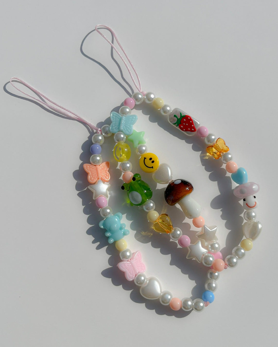 A studio shot of Buttercup Studio's Mushroom Forest Wonderland Phone Strap on top of the Froggie Friends Wonderland Phone Strap. Made with pearls, white star and heart beads, colourful beads, a blue bear bead, pink and orange butterfly beads, a smiley face bead and a special brown mushroom lampwork glass bead.