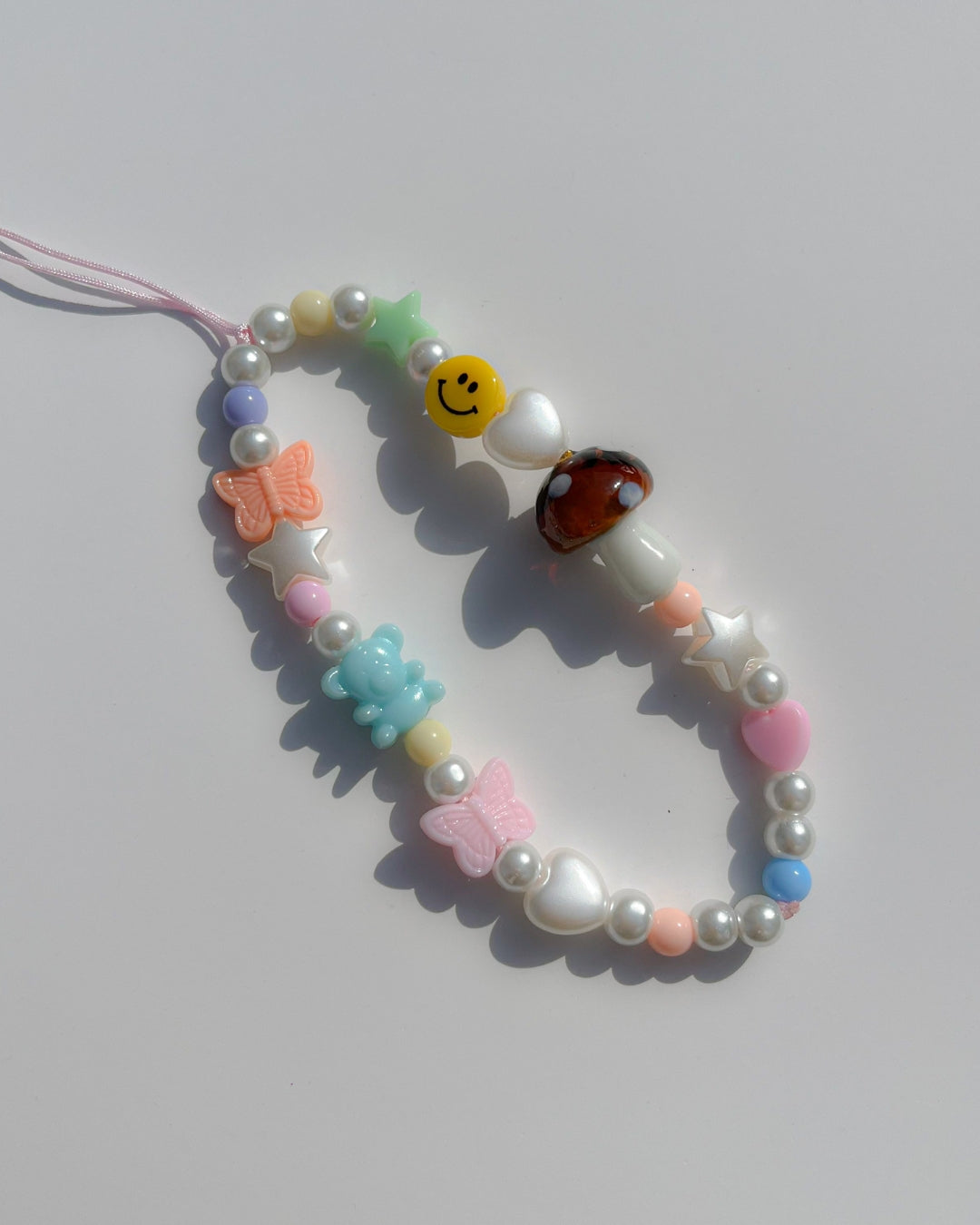 A studio shot of Buttercup Studio's Mushroom Forest Wonderland Phone Strap. Made with pearls, white star and heart beads, colourful beads, a blue bear bead, pink and orange butterfly beads, a smiley face bead and a special brown mushroom lampwork glass bead.