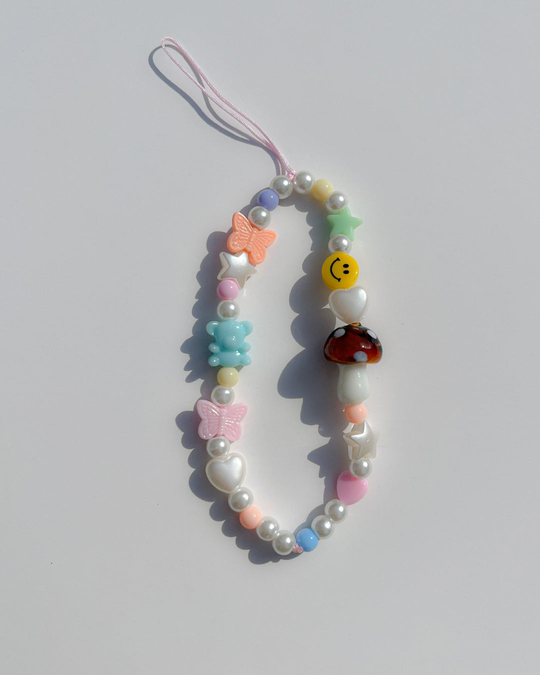 A studio shot of Buttercup Studio's Mushroom Forest Wonderland Phone Strap. Made with pearls, white star and heart beads, colourful beads, a blue bear bead, pink and orange butterfly beads, a smiley face bead and a special brown mushroom lampwork glass bead. 