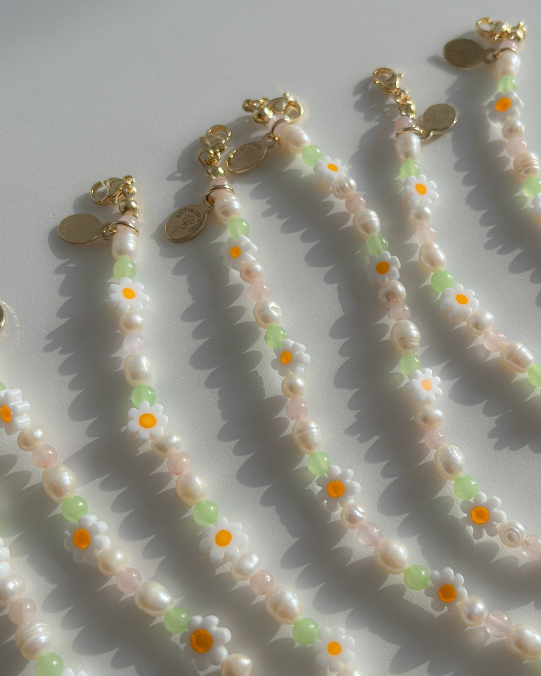 Close up studio shot of multiple Daisy For You Freshwater Pearls Bracelets by Buttercup Studio. Features freshwater pearls in assorted colours and sizes, green beads, daisy charms and a 14k gold Buttercup Studio signature oval emblem.