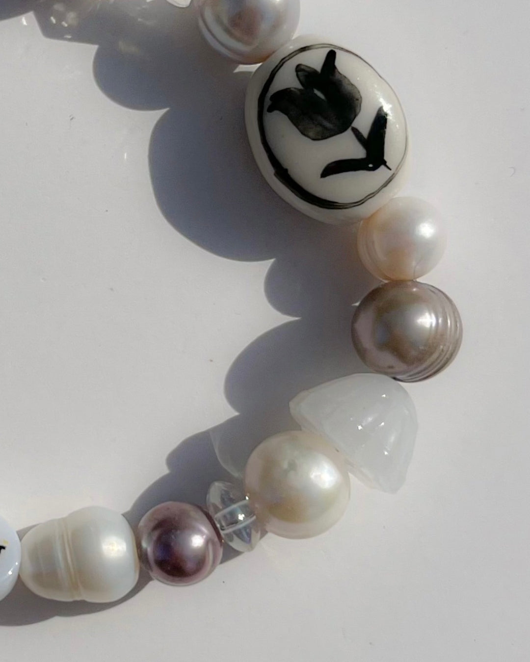 A close up studio shot of Buttercup Studio's Painter Tulip Freshwater Pearls Bracelet. Made with assorted freshwater pearls, clear and white beads, a white smiley face bead and a special lampwork white glass bead with a black hand-painted tulip.