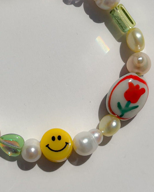 A close up studio shot of Buttercup Studio's Painting Tulip Freshwater Pearls Bracelet. Made with assorted freshwater pearls, sheer pink and green beads, a yellow smiley face bead and a special lampwork white glass bead with a red hand painted tulip.