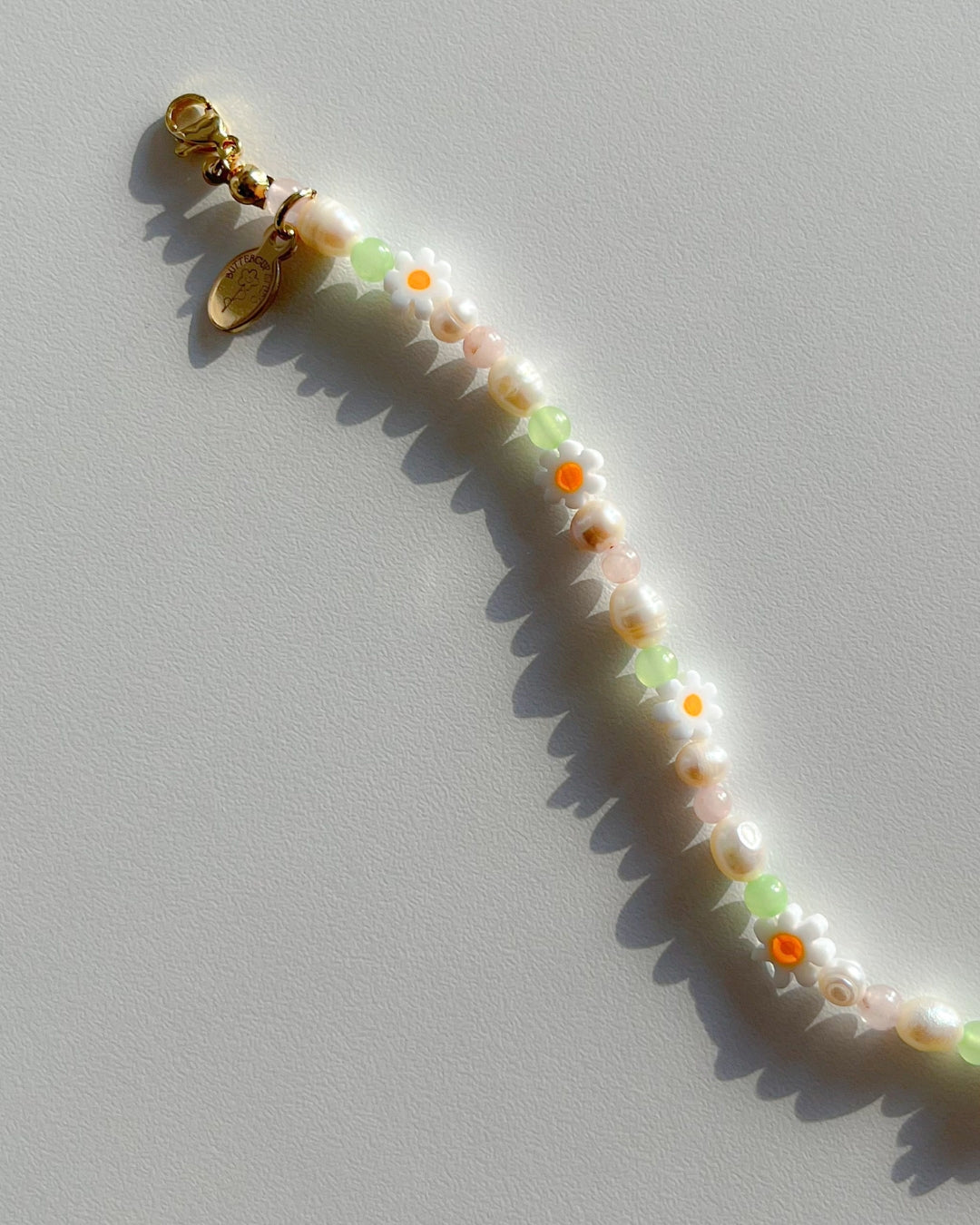 Close up studio shot of the Daisy For You Freshwater Pearls Bracelets by Buttercup Studio. Features freshwater pearls in assorted colours and sizes, green beads, daisy charms and a 14k gold Buttercup Studio signature oval emblem.