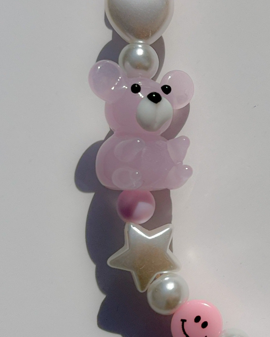A close up studio shot of Buttercup Studio's Pink Teddy Princess Phone Strap. Made with pearls, white star and heart beads, a pink butterfly bead, a pink dice bead, a pink smiley face bead and a lampwork glass pink teddy bead.