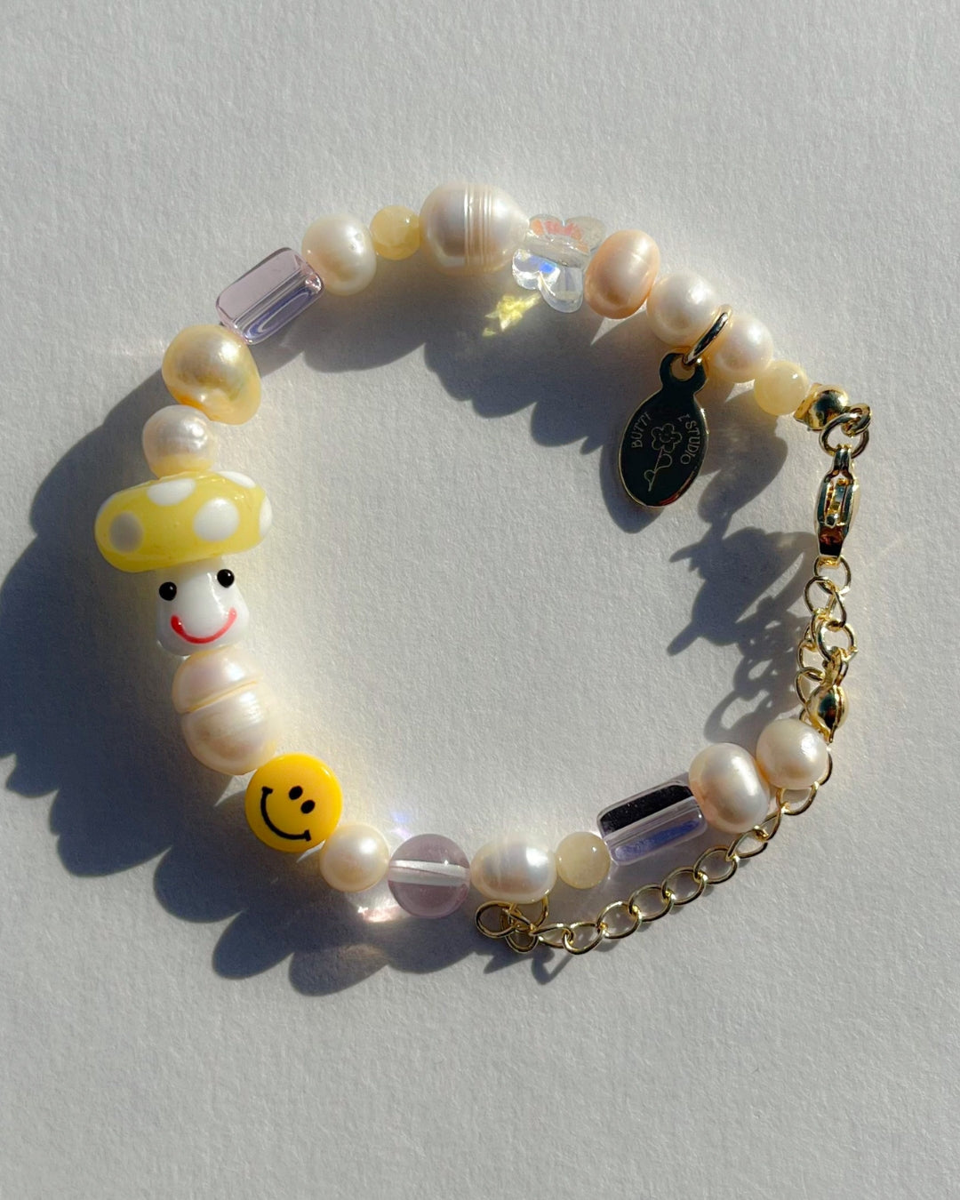 A studio shot of the Buttercup Studio Yellow Shroomie Freshwater Pearls Bracelet. Made with assorted freshwater pearls, clear and pink beads, a yellow smiley face bead and a special lampwork glass yellow mushroom bead.