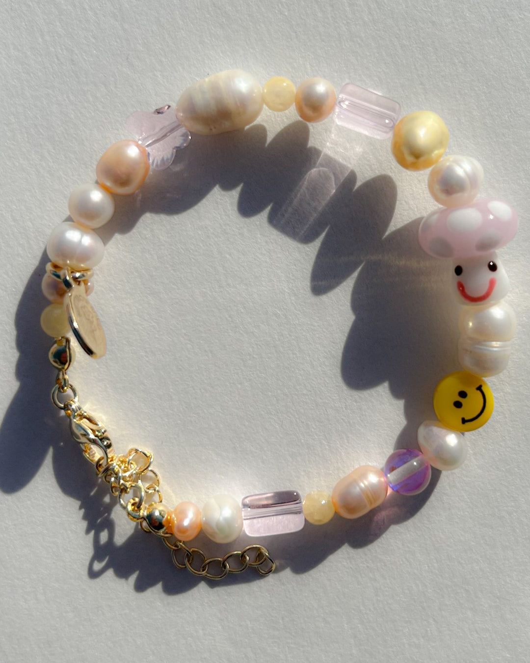 A studio shot of Buttercup Studio's Pink Shroomie Freshwater Pearls Bracelet. Includes assorted freshwater pearls, sheer pink beads, a yellow smiley face bead and a special lampwork glass pink mushroom bead.