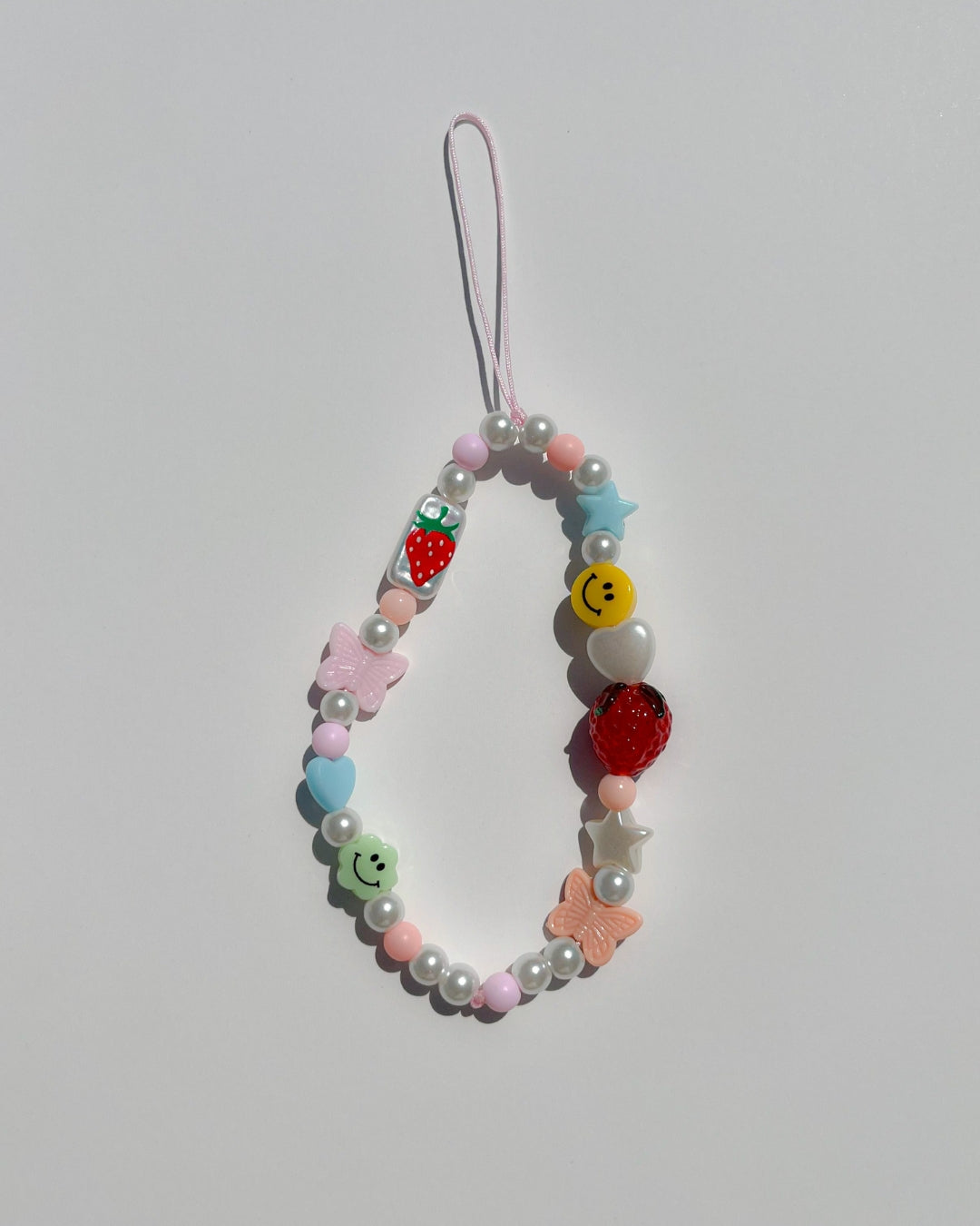 A studio shot of Buttercup Studio's Strawberry Jam Phone Strap. Made with pearls, colourful beads, white heart and star beads, a pink butterfly bead, two smiley face beads, a glass hand painted rectangle bead with a strawberry on it and a lampwork glass strawberry bead.