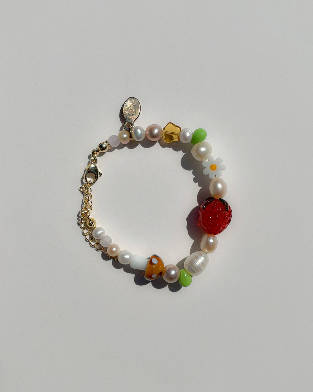A studio shot of Buttercup Studio's Summertime Strawberry Freshwater Pearls Bracelet. Features assorted freshwater pearls, green and amber beads, a white daisy bead, a brown lampwork mushroom glass bead and a lampwork glass strawberry bead. 