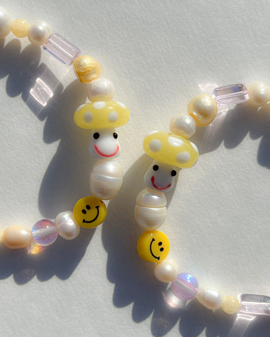 A studio shot of two Buttercup Studio Yellow Shroomie Freshwater Pearls Bracelets. Made with assorted freshwater pearls, clear and pink beads, a yellow smiley face bead and a special lampwork glass yellow mushroom bead.