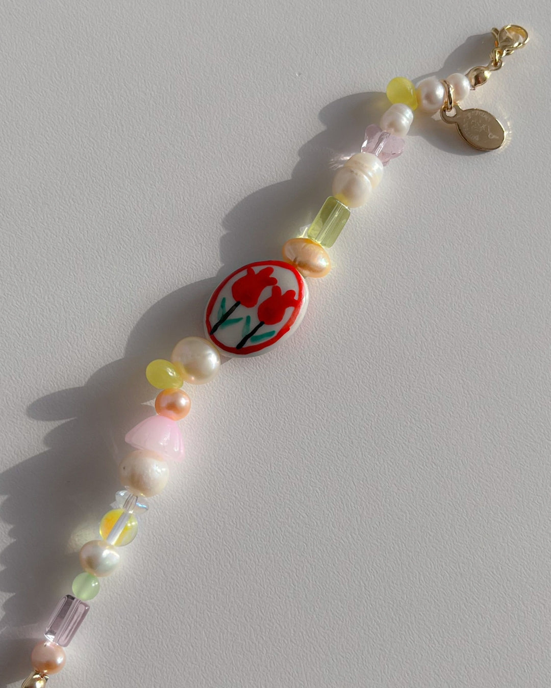 A close up studio shot of Buttercup Studio's Tulip Field Freshwater Pearls Bracelet. Made with assorted freshwater pearls, pink and yellow beads and a lampwork hand painted glass bead with two red tulips.