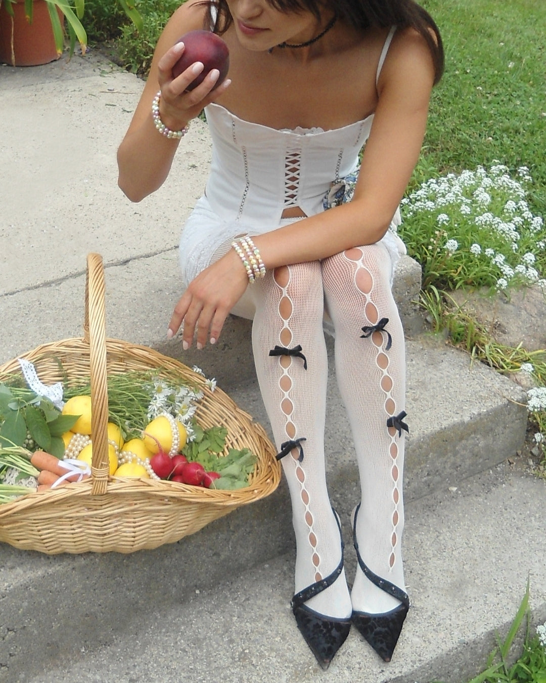 A close up shot of a model sitting on concrete steps, wearing a white spaghetti strap corset and white netted stockings with black bows throughout. They are sniffinf a peach in one hand while the other hand is reaching for a woven picnic basket filled with flowers, lemons, radishes, herbs and strong pearls. The model wears Buttercup Studio's Timeless Pastel Freshwater Pearls Bracelet on their wrists.