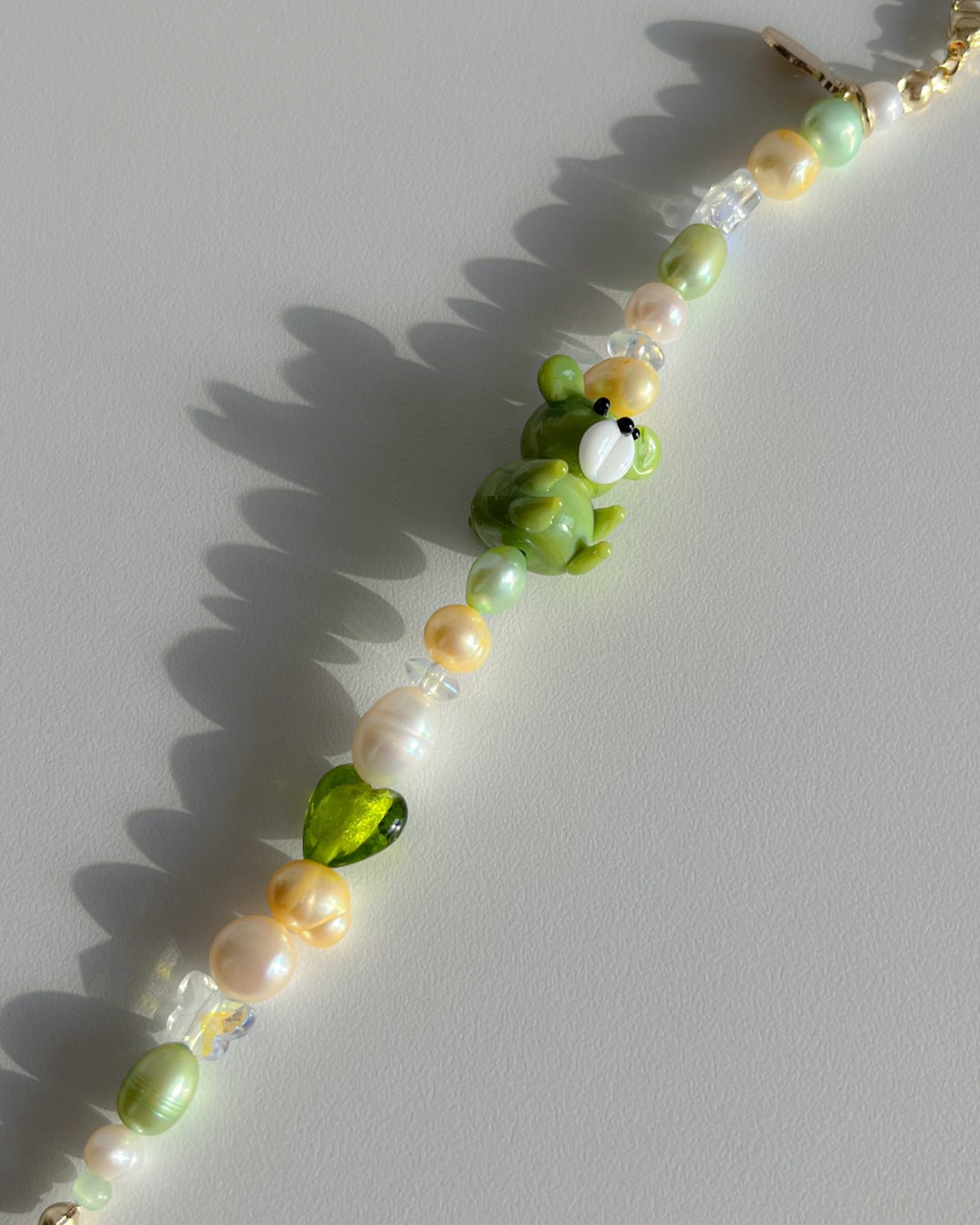 A close up studio shot of Buttercup Studio's Forest Teddy Freshwater Pearls Bracelet. Features clear beads, a sheer green heart bead, assorted yellow, green and white freshwater pearls, and a special green teddy lampwork glass bead.