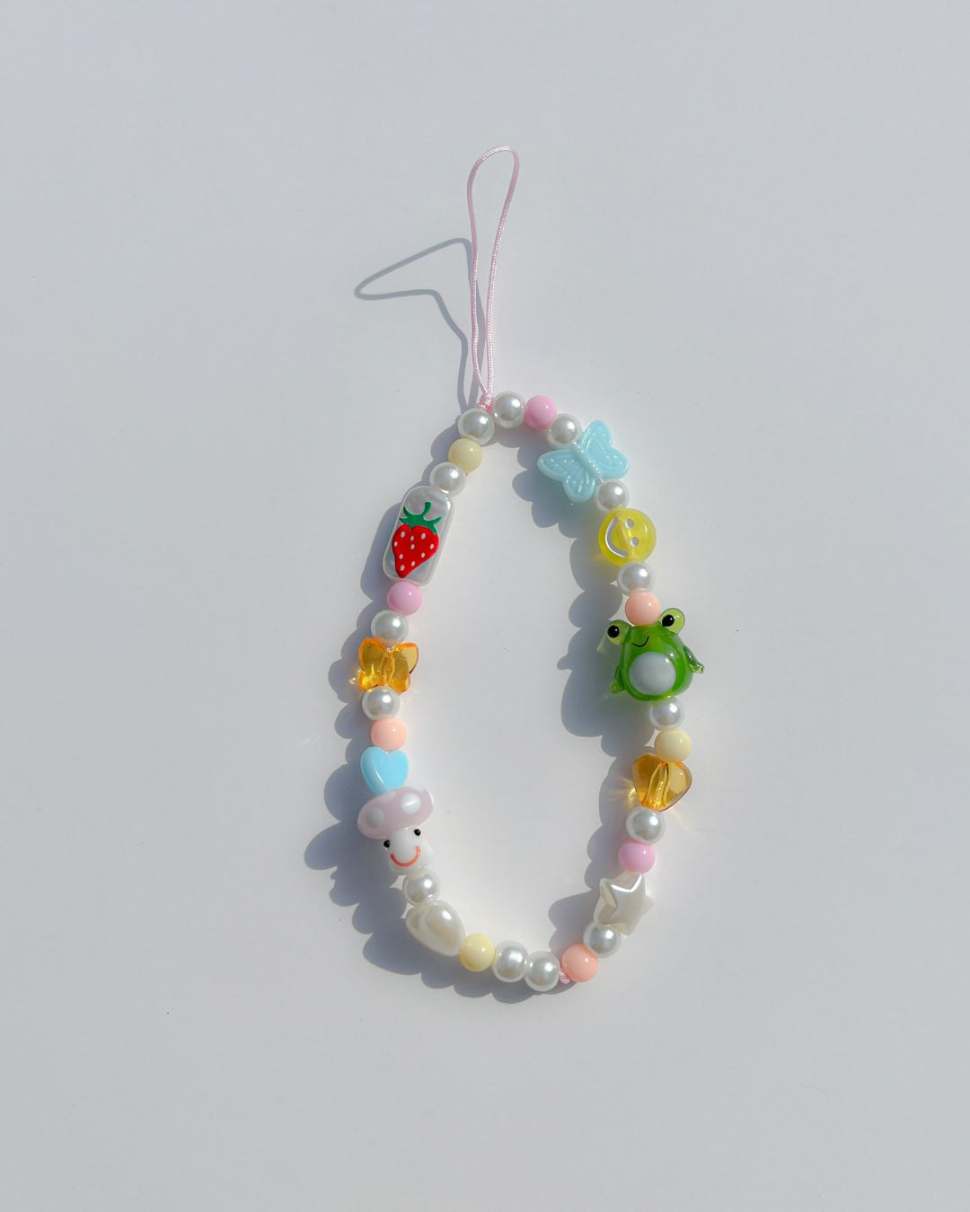 A studio shot of Buttercup Studio's Froggie Friends Wonderland Phone Strap. Made with pink, yellow and coral colourful beads, a blue butterfly bead, a yellow smiley face bead, amber heart and butterfly beads, blue and white heart and star beads, a handpainted strawberry rectangle bead, a special mushroom lampwork glass bead, a special green frog lampwork glass bead and pearls.