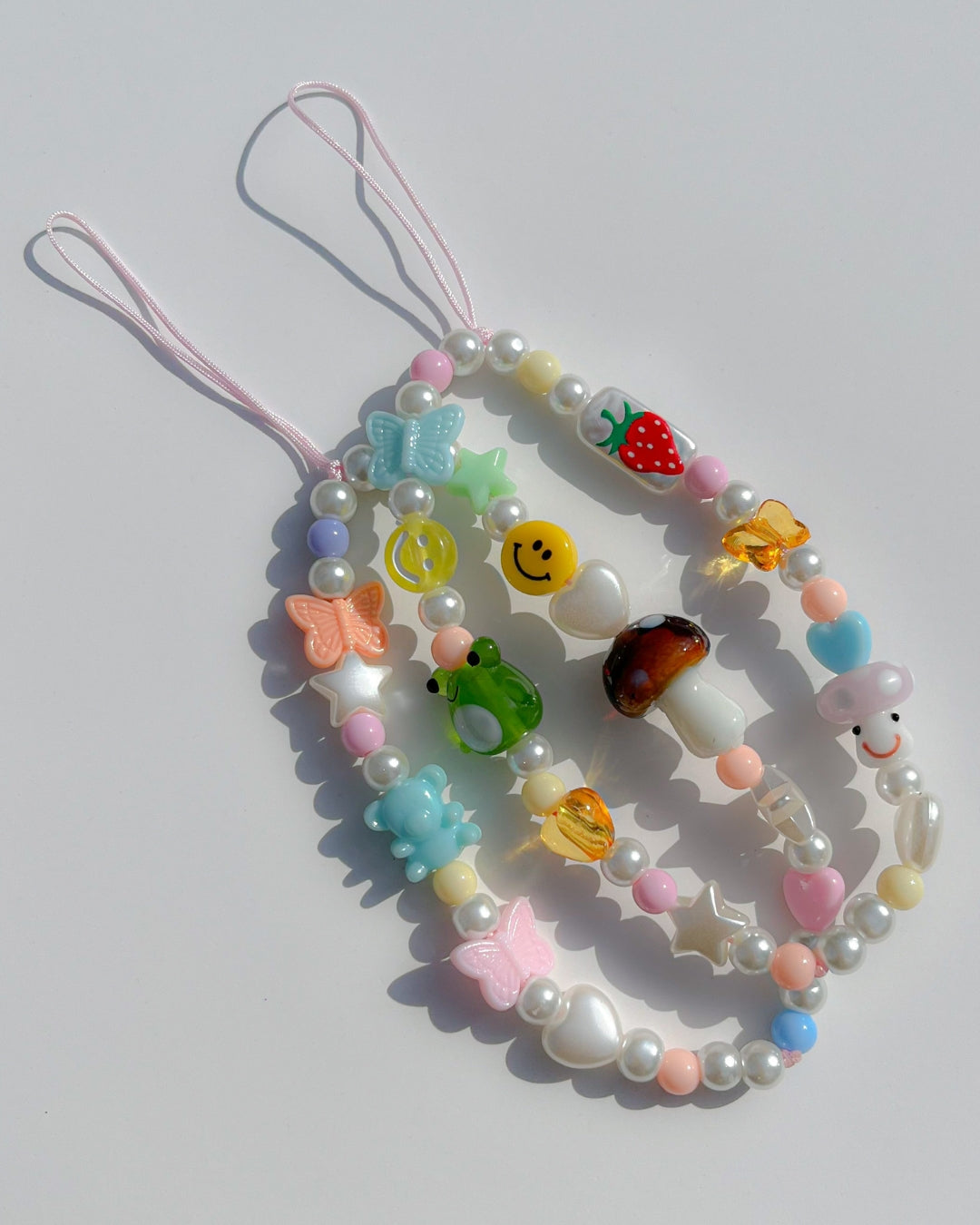 A studio shot of Buttercup Studio's Froggie Friends Wonderland Phone Strap on top of Butercup Studio's Mushroom Forest Wonderland Phone Strap. Made with pink, yellow and coral colourful beads, a blue butterfly bead, a yellow smiley face bead, amber heart and butterfly beads, blue and white heart and star beads, a handpainted strawberry rectangle bead, a special mushroom lampwork glass bead, a special green frog lampwork glass bead and pearls.