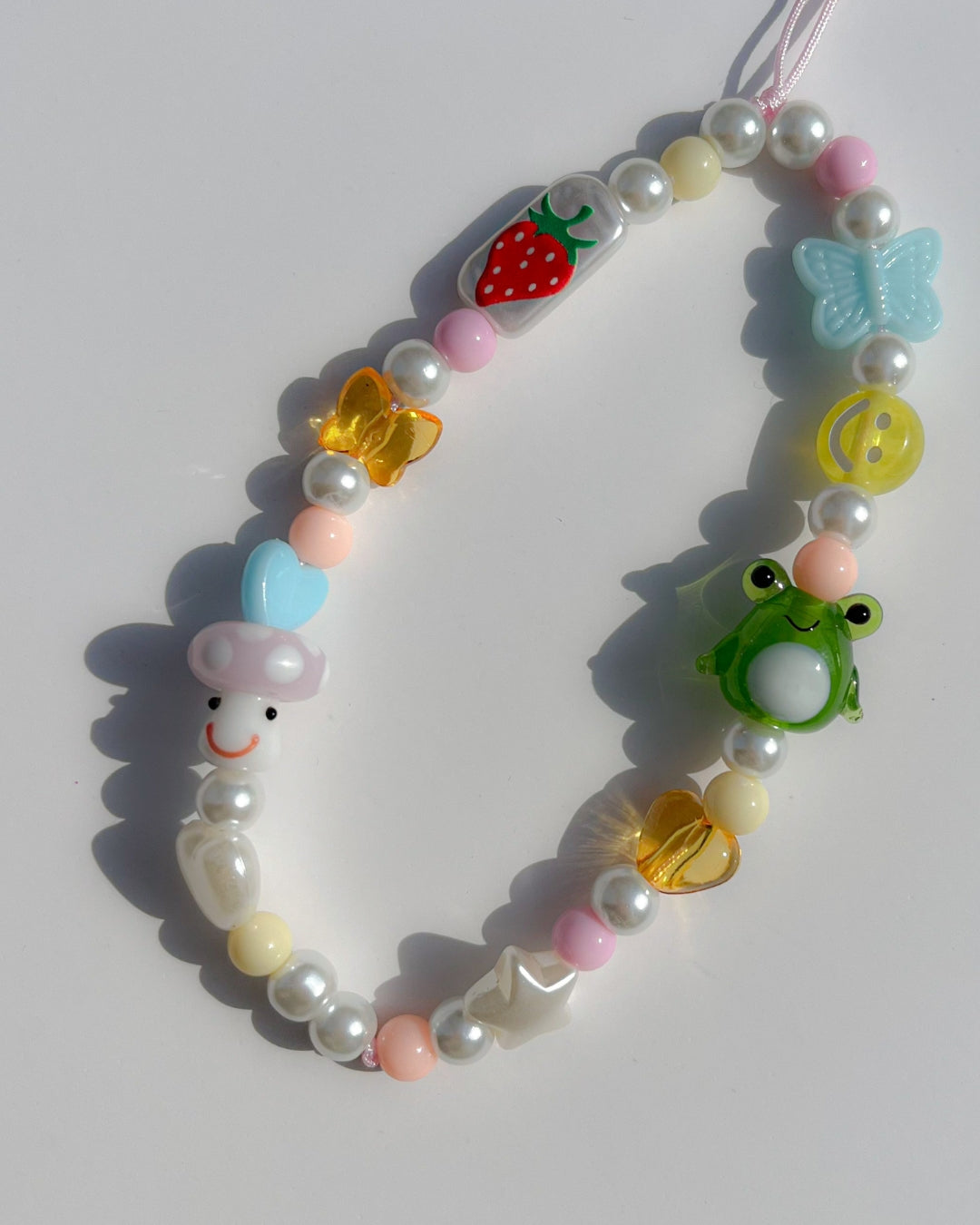 A studio shot of Buttercup Studio's Froggie Friends Wonderland Phone Strap. Made with pink, yellow and coral colourful beads, a blue butterfly bead, a yellow smiley face bead, amber heart and butterfly beads, blue and white heart and star beads, a handpainted strawberry rectangle bead, a special mushroom lampwork glass bead, a special green frog lampwork glass bead and pearls.