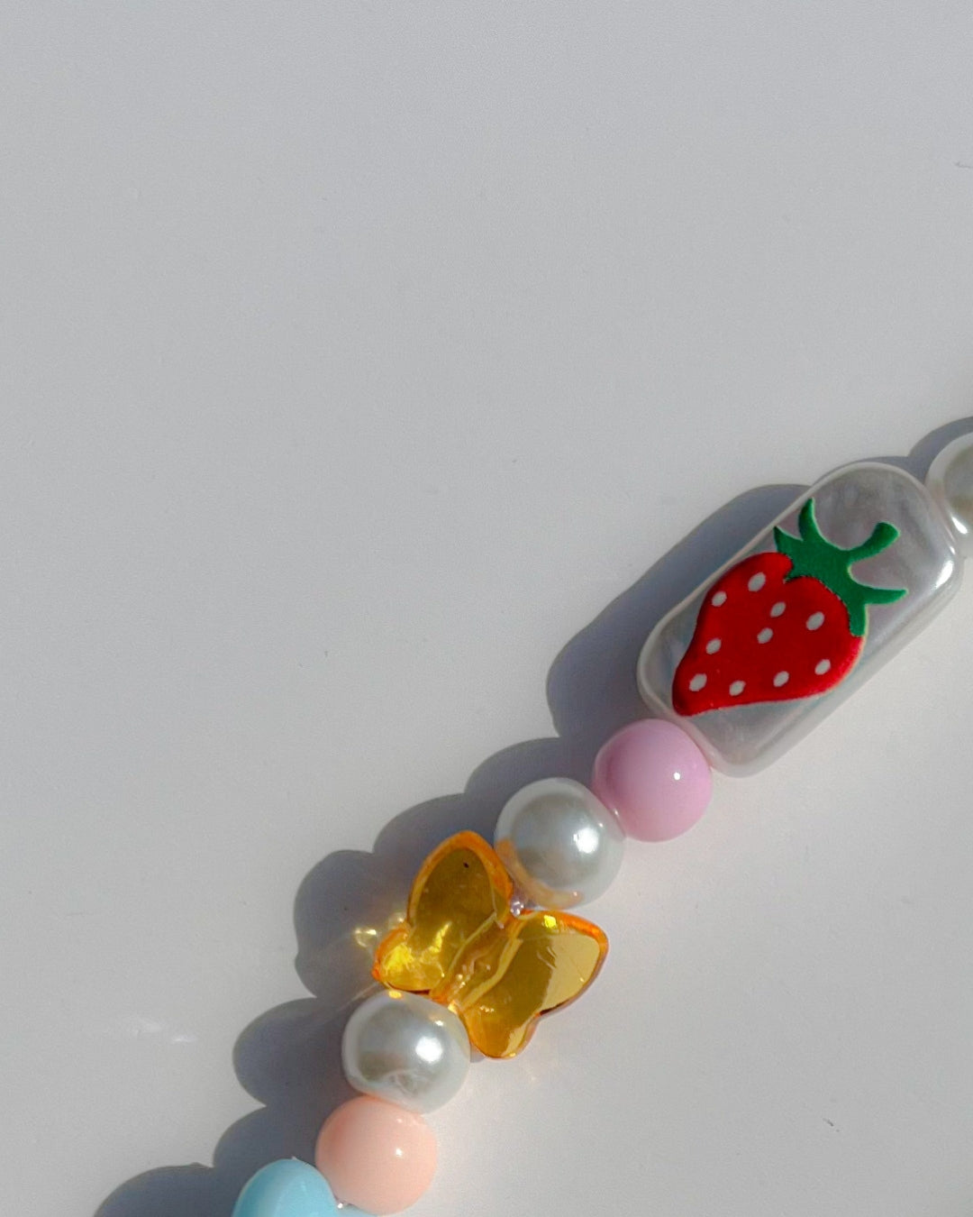 A close up studio shot of Buttercup Studio's Froggie Friends Wonderland Phone Strap. Made with a handpainted strawberry rectable bead, an amber butterfly bead and white, pink, coral and blue beads and pearls.