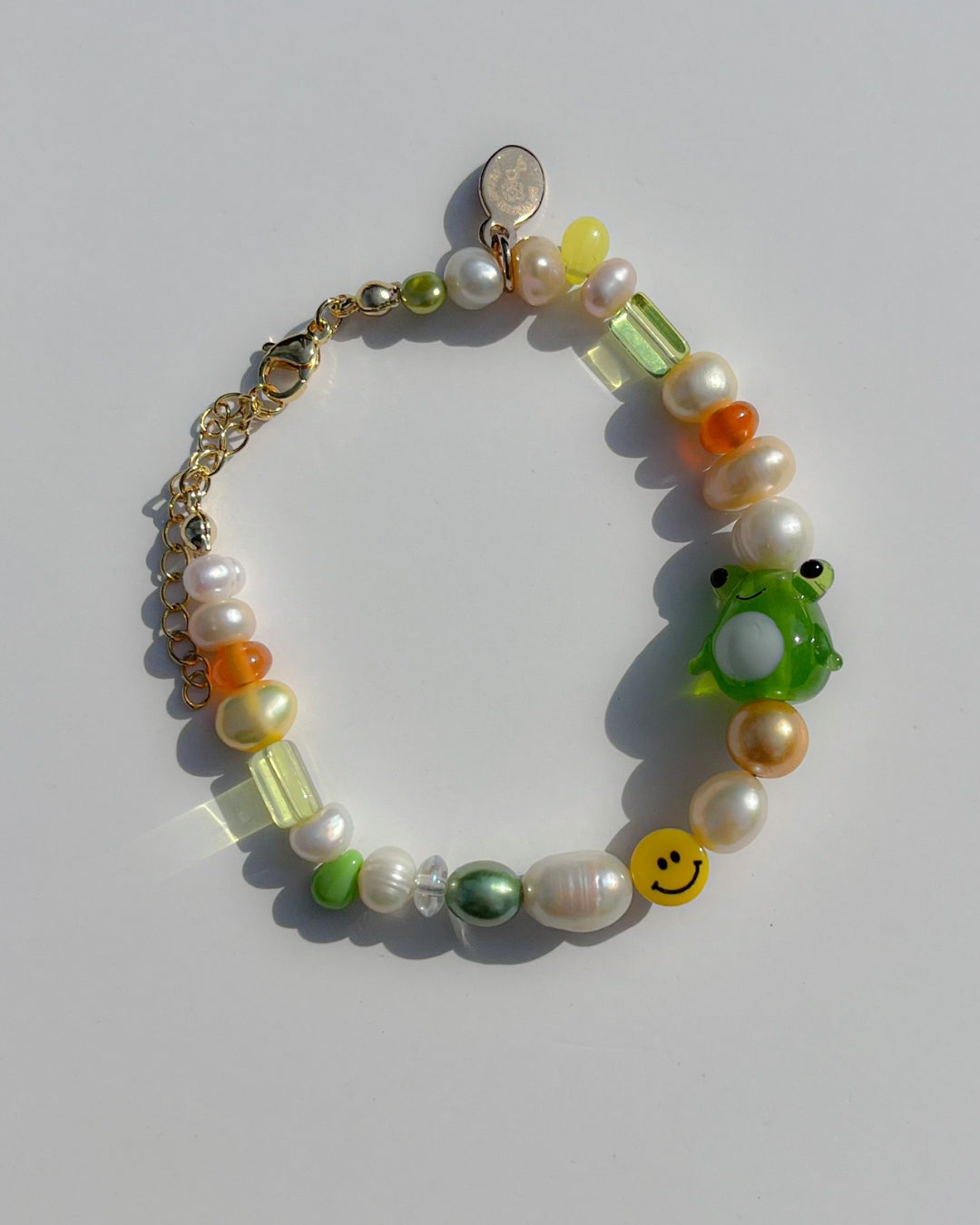 A studio shot of Buttercup Studio's Happy Froggie Freshwater Pearls Bracelet. Made with assorted freshwater pearls, sheer amber and green beads, a yellow smiley face bead and a special green frog lampwork glass bead.