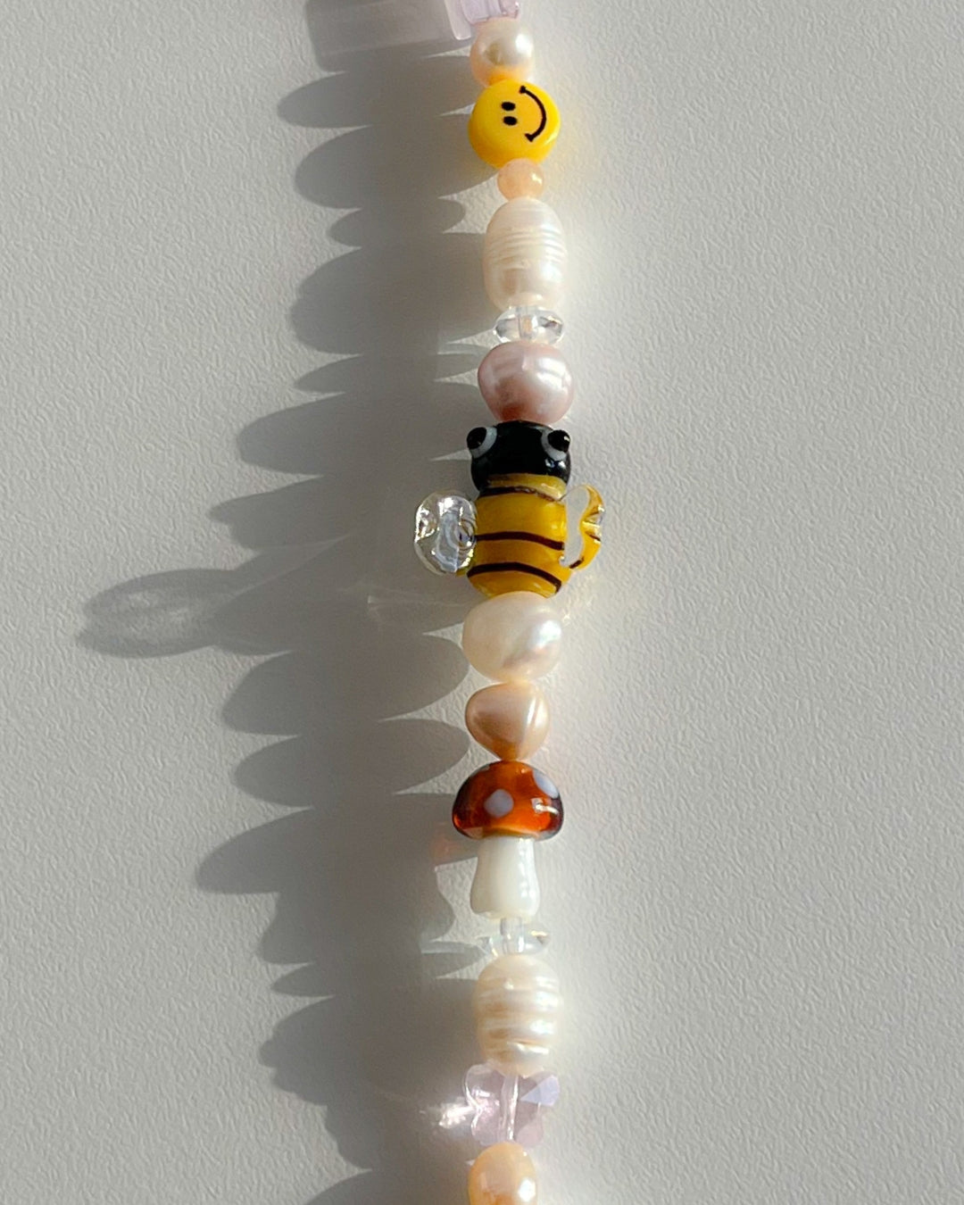 A close up studio shot of Buttercup Studio's Honeycomb Bee Freshwater Pearls Bracelet. Made with assorted freshwater pearls, clear pink and amber beads, a yellow smiley face bead, a special brown mushroom lampwork glass bead and a special yellow bee lampwork glass bead.