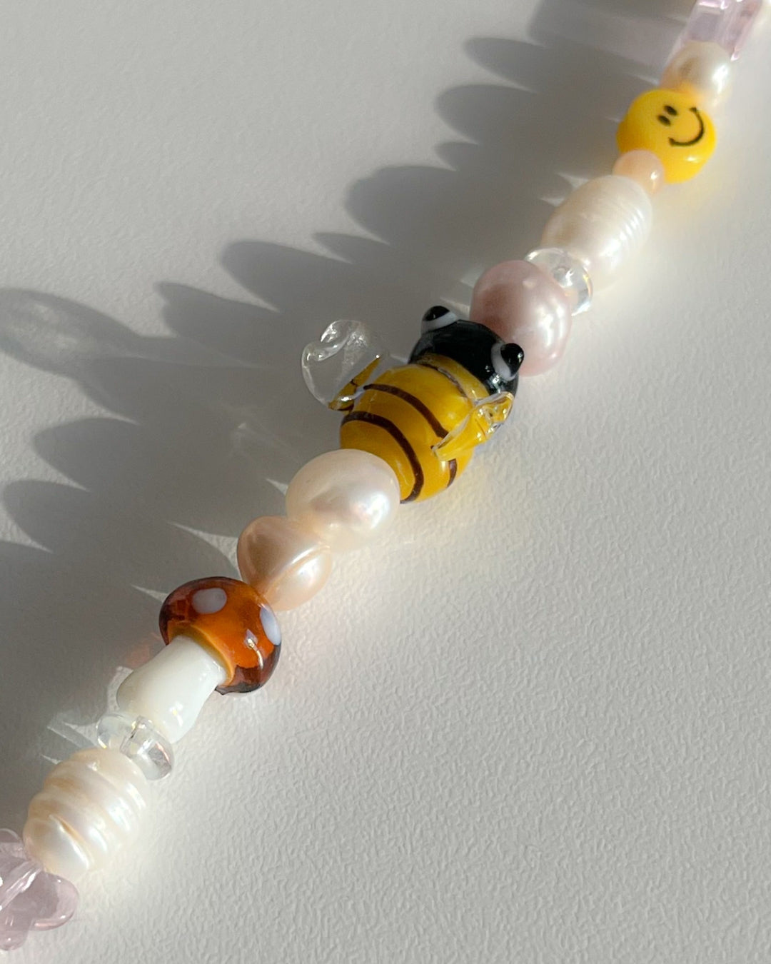 A studio shot of Buttercup Studio's Honeycomb Bee Freshwater Pearls Bracelet. Made with assorted freshwater pearls, clear pink and amber beads, a yellow smiley face bead, a special brown mushroom lampwork glass bead and a special yellow bee lampwork glass bead. 