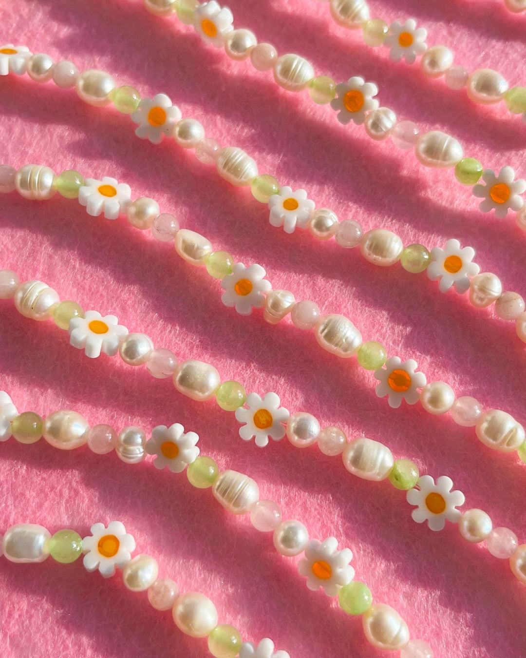 Close up pink background studio shot of multiple Daisy For You Freshwater Pearls Bracelets by Buttercup Studio. Features freshwater pearls in assorted colours and sizes, green beads and daisy charms.