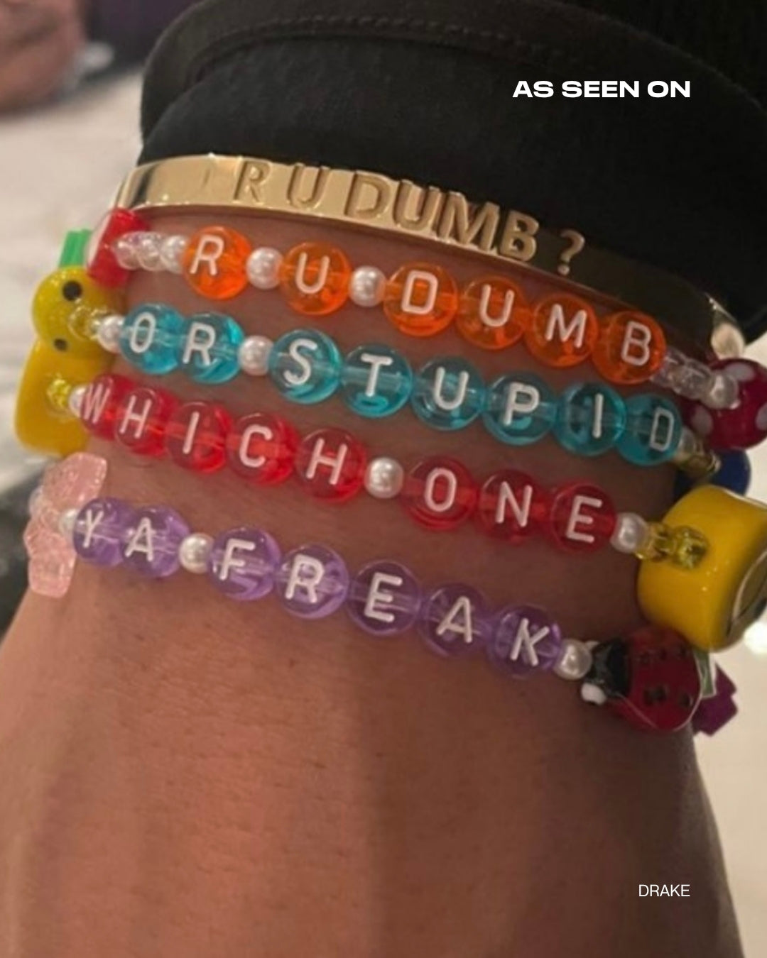 A close up of Buttercup Studio's Aubrey Set on Drake's wrist. A gold curved bracelet band carved with the phrase R U DUMB? with the Drake Custom Bracelet set in orange beads spelling out R U DUMB? below it, and a Drake custom bracelet set in teal beads spelling out OR STUPID below that, and a Drake Custom Bracelet in red beads spelling out WHICH ONE under it, and a Drake Custom Bracelet set in purple beads spelling out YA FREAK at the bottom. Image credits: @champagnepapi on Instagram.