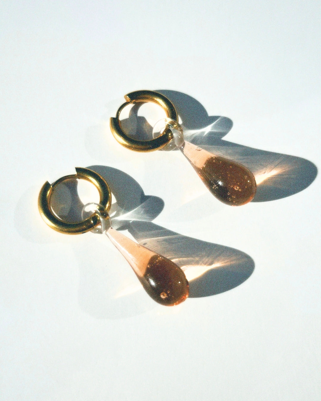 A studio shot of the Celeste earrings in coco brown from Buttercup Studio. The Celeste Earrings are made with thick 18k gold plated tarnish-resistant, hypoallergenic 12 mm thick hoops and high-quality 22mm lampwork glass water drop pendants.